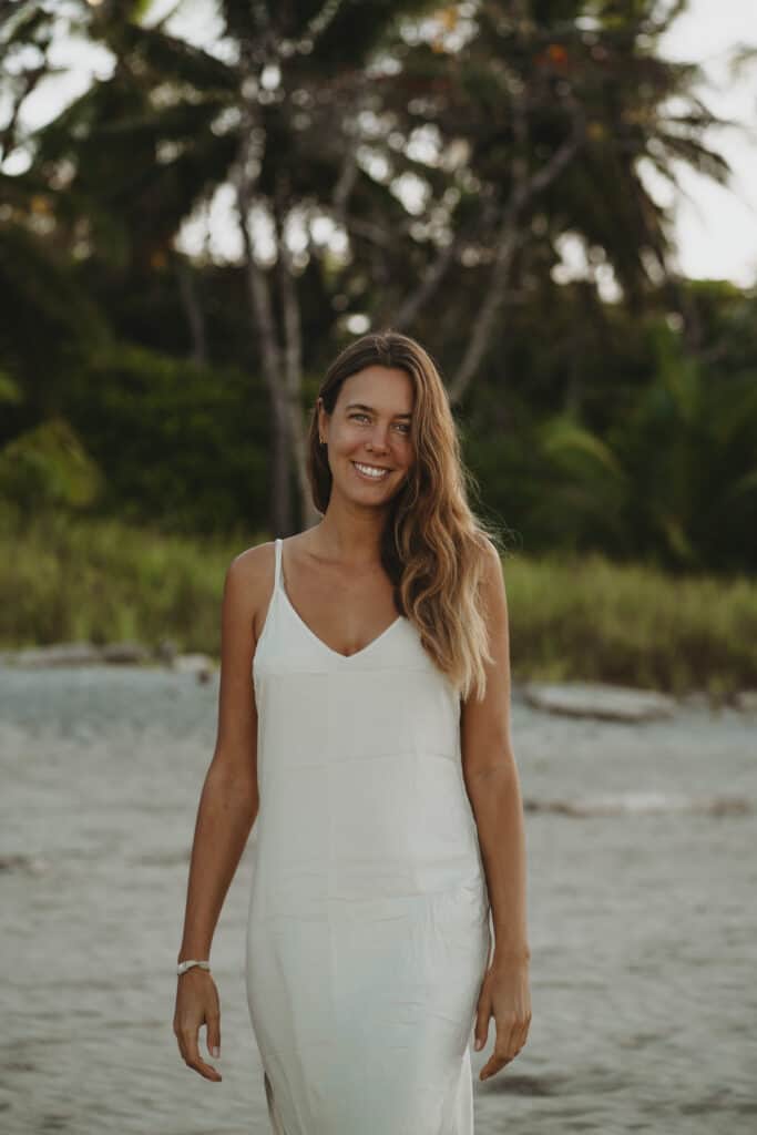 A woman with long hair in a white dress stands on a sandy beach with lush greenery and tall palm trees in the background. She is smiling and appears relaxed in the natural, serene setting.